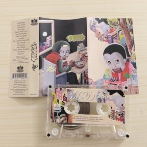 Op de afbeelding: Een transparante cassetteband met een kleurrijke albumhoes. De illustratie toont cartoon illustraties en het woord "OASIS" in graffiti stijl. De cassette is open en toont de illustratie en de tracklist.