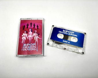 May include: A clear cassette tape and case with the text "K-POP DEMON HUNTERS". The cassette case has a red cover with illustrations of three figures and silhouettes. The cassette tape has a blue label with white text.