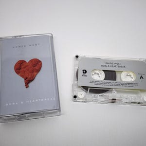 Puede incluir: Una cinta de cassette transparente y su estuche con el álbum "808s & Heartbreak" de Kanye West. El estuche tiene un corazón rojo. La cinta de cassette es visible junto al estuche.