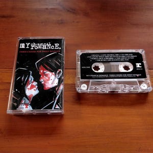 May include: A clear cassette tape and case featuring the album "Three Cheers for Sweet Revenge" by My Chemical Romance. The case has a black and red graphic of two figures. The cassette tape has song titles printed on it.