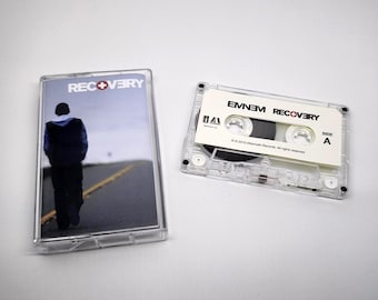 Könnte beinhalten: Eine transparente Kassette und Hülle mit dem Album "Recovery" von Eminem. Die Hülle zeigt eine Person, die eine Straße entlanggeht. Auf der Kassette sind der Albumtitel und der Künstlername aufgedruckt.