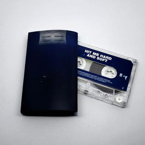 May include: A dark blue cassette tape case and a clear cassette tape with a dark blue label. The label reads "HIT ME HARD AND SOFT" in white text. The cassette tape is partially out of the case.