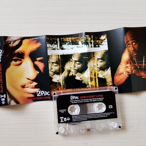 May include: A 2Pac Greatest Hits cassette tape with its cover art unfolded. The cover features portraits of 2Pac, with the cassette itself visible. The cassette case is clear, and the album art is visible.