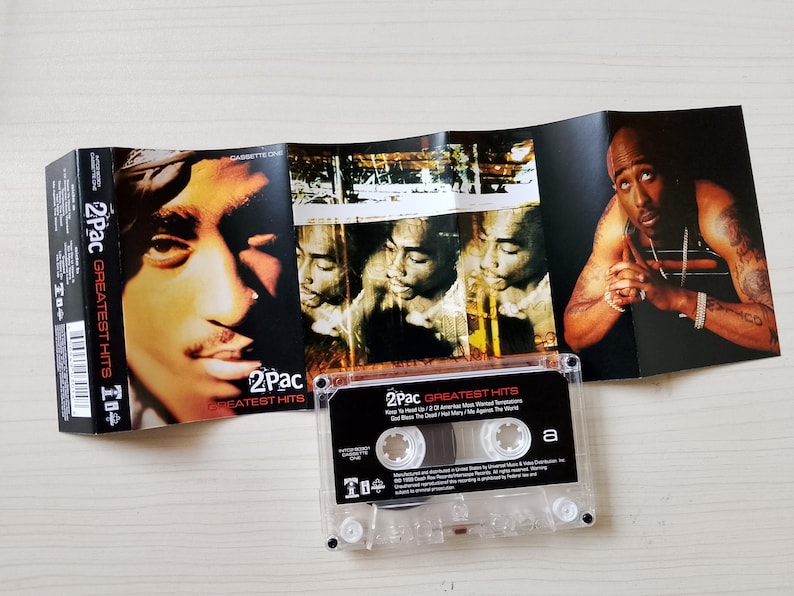 May include: A 2Pac Greatest Hits cassette tape with its cover art unfolded. The cover features portraits of 2Pac, with the cassette itself visible. The cassette case is clear, and the album art is visible.