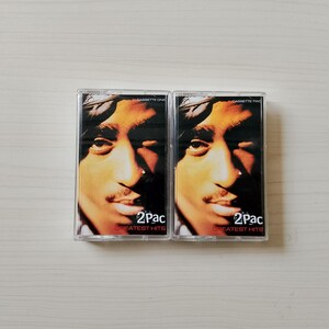 May include: Two cassette tapes featuring the album cover of 2Pac's Greatest Hits. The album art shows a close-up of 2Pac's face with a gold and brown color scheme. The text "2Pac" and "Greatest Hits" are visible.