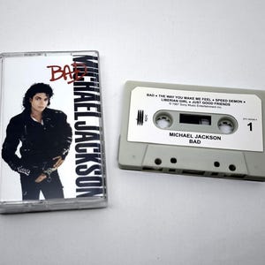Op de afbeelding: Een vintage cassettebandje en hoesje met het album "Bad" van Michael Jackson. De hoes heeft een foto van Jackson in een zwart jasje. De cassette is grijs met de albumtitel en tracklist erop gedrukt.