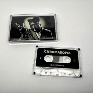 May include: A clear cassette tape with a black label that reads "CHROMAKOPIA" and "TYLER, THE CREATOR." The cassette case contains a black and white image of a man. The cassette is next to its case on a white surface.