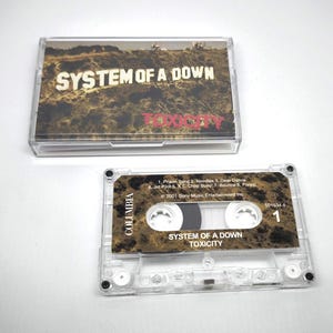 May include: A clear cassette tape and case featuring the album "Toxicity" by System of a Down. The cassette tape has a brown and gold label with the band's name and album title. The case displays the album title and band name.