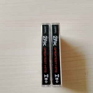 May include: Two cassette tapes of 2Pac's Greatest Hits. The cassette cases are clear with black and red text. The text includes "2Pac" and "Greatest Hits". The tapes are standing upright on a light-colored surface.