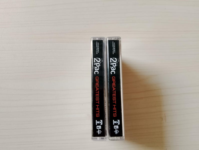 May include: Two cassette tapes of 2Pac's Greatest Hits. The cassette cases are clear with black and red text. The text includes "2Pac" and "Greatest Hits". The tapes are standing upright on a light-colored surface.