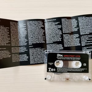 May include: A clear cassette tape labeled "2Pac Greatest Hits" with the tracklist visible. The cassette is next to a black and white printed insert with song lyrics and credits. The cassette tape is on a light-colored surface.