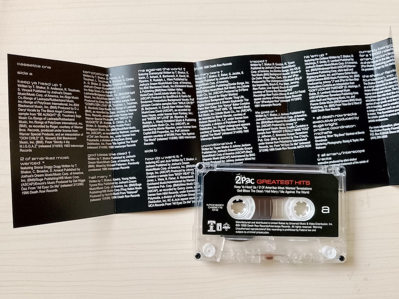 May include: A clear cassette tape labeled "2Pac Greatest Hits" with the tracklist visible. The cassette is next to a black and white printed insert with song lyrics and credits. The cassette tape is on a light-colored surface.