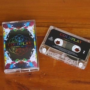 May include: A clear cassette tape and case featuring the album "A Head Full of Dreams" by Coldplay. The cassette case has a colorful design with the band's name and album title. The cassette tape is visible.