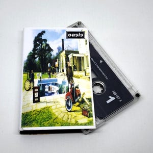 Op de afbeelding: Een cassettebandje met de albumhoes van Oasis. De hoes toont een rode scooter, een vintage klok en een gebouw. Het Oasis-logo staat rechtsboven. De cassette is zwart met witte tekst.