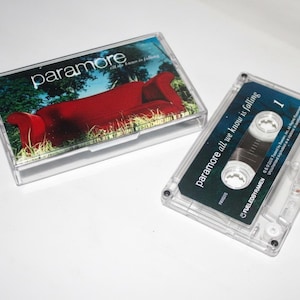 May include: A clear cassette tape and case featuring the band Paramore. The case has an image of a red couch in a field. The cassette tape is labeled "paramore all we know is falling."