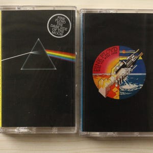 Puede incluir: Dos cintas de cassette vintage. La primera presenta una portada negra con un prisma refractando la luz en un arcoíris, y el texto "PINK FLOYD THE DARK SIDE OF THE MOON". La segunda cinta tiene una portada negra con un gráfico de una mano, una puesta de sol y el texto "PINK FLOYD WISH YOU WERE HERE".