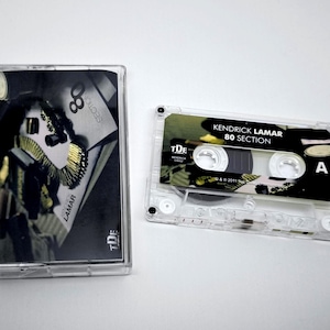 May include: A clear cassette tape and its case are displayed. The cassette case features a black and white image with gold accents and the name "LAMAR". The cassette tape has the text "KENDRICK LAMAR 80 SECTION" and the letter "A".