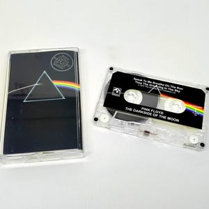 Può includere: Una musicassetta dei Pink Floyd con l'album "The Dark Side of the Moon". La custodia nera presenta un design a prisma e arcobaleno. La cassetta è trasparente con un'etichetta nera e arcobaleno.