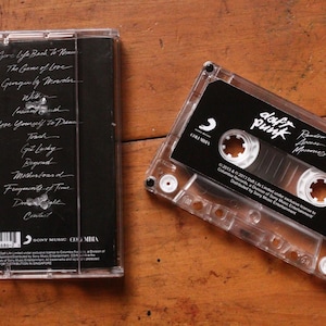 May include: A clear cassette tape with a black label featuring the band name "Daft Punk" and album title. The cassette case is next to its cover, which lists song titles in white text. The background is a wooden surface.