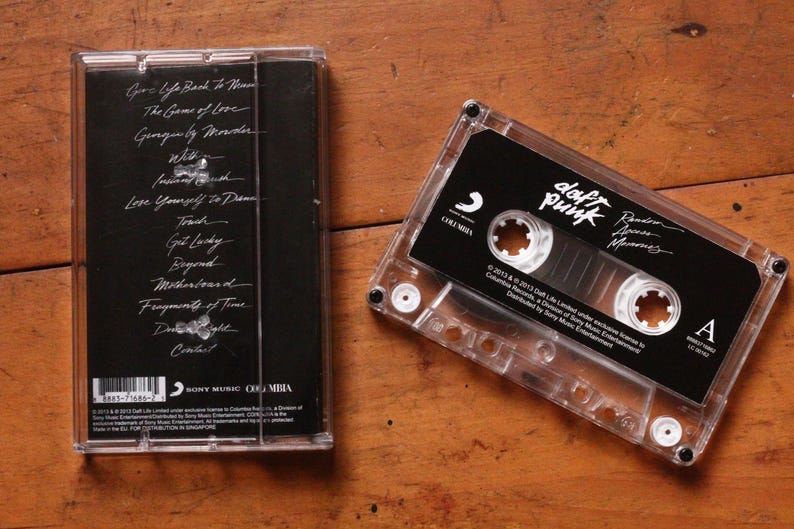 May include: A clear cassette tape with a black label featuring the band name "Daft Punk" and album title. The cassette case is next to its cover, which lists song titles in white text. The background is a wooden surface.