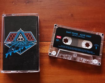 May include: Two cassette tapes on a wooden surface. One cassette case is black with a blue and white graphic design. The other cassette tape is clear with a black label that reads "DAFT PUNK ALIVE 2007".