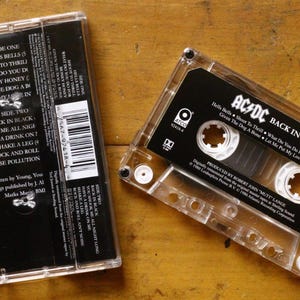 May include: A clear cassette tape with a black label featuring the text "AC/DC BACK IN BLACK" and a tracklist. The cassette case is transparent, and the image is set against a wooden surface. The album's tracklist is visible.