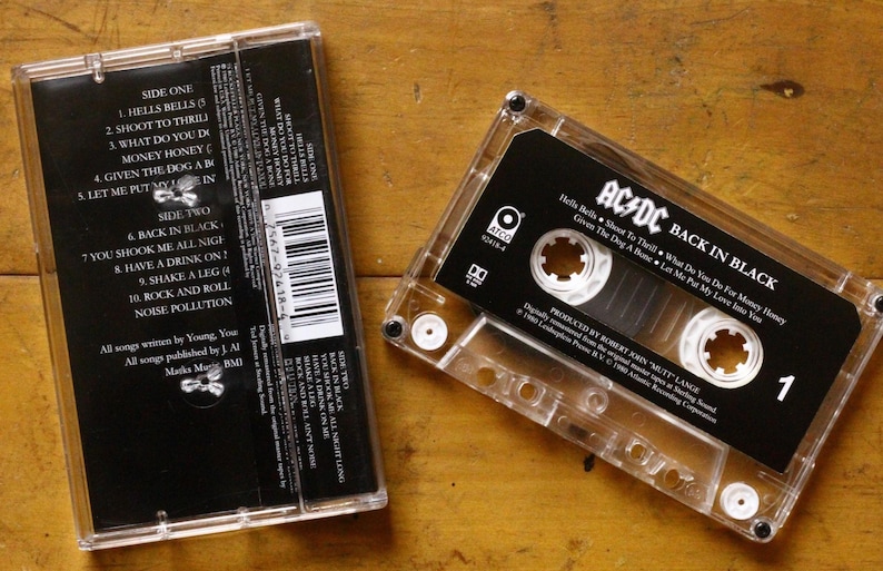 May include: A clear cassette tape with a black label featuring the text "AC/DC BACK IN BLACK" and a tracklist. The cassette case is transparent, and the image is set against a wooden surface. The album's tracklist is visible.