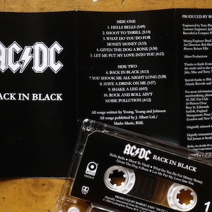 May include: AC/DC's "Back in Black" album on cassette tape. The black cassette case is open, revealing the tracklist and album art. The album title and band logo are prominently displayed.