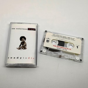 Op de afbeelding: Een transparante cassetteband en hoesje met het album "Ready to Die" van The Notorious B.I.G. De albumhoes toont een baby. De cassetteband is gelabeld met de albumtitel en artiest.
