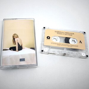 Op de afbeelding: Een doorzichtig plastic cassettehoesje met een albumhoes waarop een persoon in een zwarte jurk op een wit oppervlak zit, naast een transparante cassette met een beige label met de tekst Sabrina Carpenter Emails I Can't Send.
