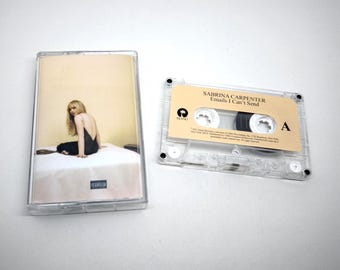 May include: A clear plastic cassette case with an album cover insert featuring a person in a black dress sitting on a white surface, alongside a transparent cassette tape with a tan label reading Sabrina Carpenter Emails I Can't Send.