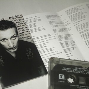 May include: A black and white photo of a man with text from a Green Day album. The image includes a cassette tape and lyrics from the album "Nimrod". The album cover features the text "Green Day / Nimrod".