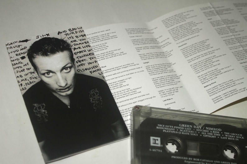 May include: A black and white photo of a man with text from a Green Day album. The image includes a cassette tape and lyrics from the album "Nimrod". The album cover features the text "Green Day / Nimrod".