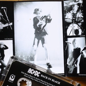 May include: Black and white image of an AC/DC "Back in Black" cassette tape and album art. The album art features band members performing live. The cassette tape is in the foreground, with the album title and song titles visible.