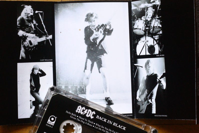 May include: Black and white image of an AC/DC "Back in Black" cassette tape and album art. The album art features band members performing live. The cassette tape is in the foreground, with the album title and song titles visible.