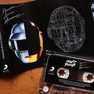 May include: A Daft Punk "Random Access Memories" cassette tape and album art. The album art features the iconic robot helmets, one in a glossy finish and the other a technical drawing. The cassette tape is clear with a black label.
