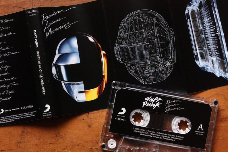 May include: A Daft Punk "Random Access Memories" cassette tape and album art. The album art features the iconic robot helmets, one in a glossy finish and the other a technical drawing. The cassette tape is clear with a black label.