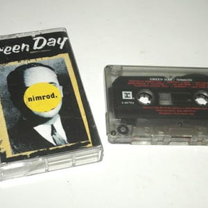 May include: A Green Day "Nimrod" cassette tape and case. The case features the band's name and album title, along with a black and white image of a man with a yellow circle over his face. The cassette tape is black with white text.