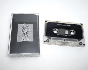 May include: A clear plastic cassette case with a black insert featuring a white pulsar wave graphic, alongside a matching clear cassette tape with a black label containing the text Joy Division Unknown Pleasures FACT 10 A Factory Cassette.