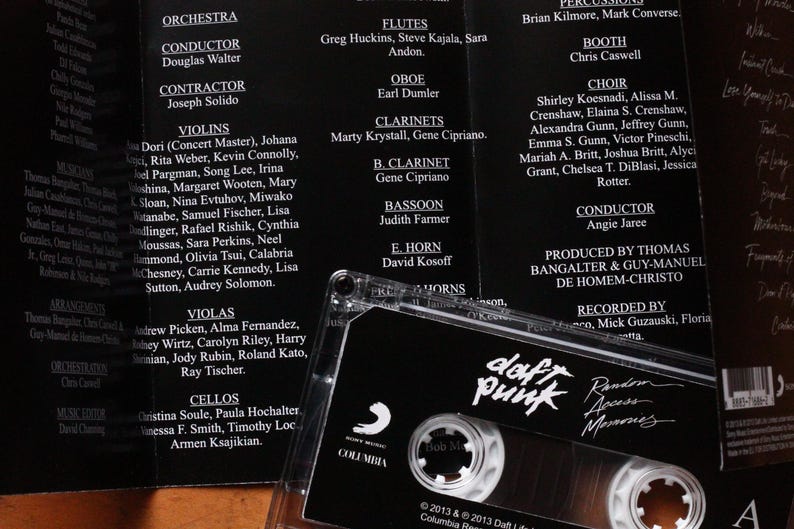 May include: A black cassette tape with the Daft Punk logo and album title "Random Access Memories" is displayed. The cassette is in a clear plastic case, with the album credits in the background.