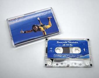 May include: A clear cassette tape with a blue and white label, featuring the text "Masayoshi Takanaka All Of Me." The cassette case shows a person skydiving. The cassette is next to its case.