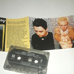 May include: A Green Day "Nimrod" cassette tape with album art featuring band members. The cassette case is transparent, and the album art includes a photo of a man with a yellow circle over his face and the band's name.