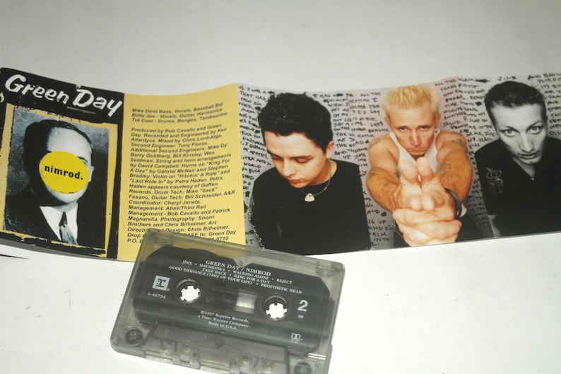 May include: A Green Day "Nimrod" cassette tape with album art featuring band members. The cassette case is transparent, and the album art includes a photo of a man with a yellow circle over his face and the band's name.