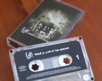 May include: A clear cassette tape featuring the album "Take a Look in the Mirror" by Korn. The cassette case is visible, with the album art in the background. The tape has the band's logo and the number 1.