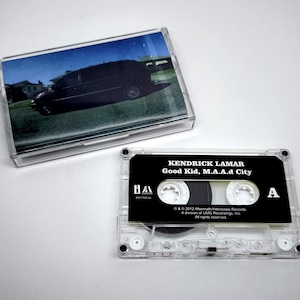 Puede incluir: Una cinta de cassette transparente con una etiqueta negra con el texto "KENDRICK LAMAR Good Kid, M.A.A.d City". La caja del cassette tiene una foto de una furgoneta negra. La cinta está en una caja de plástico transparente.