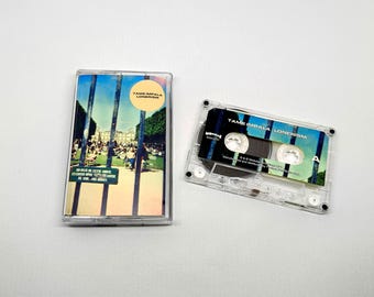 May include: A clear cassette tape and case featuring the album "Lonerism" by Tame Impala. The case has a colorful image of a park scene with text. The cassette tape is next to the case.