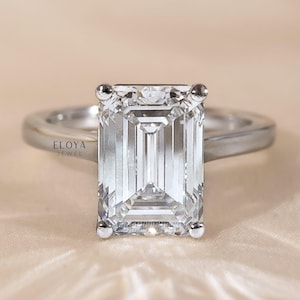 May include: A silver ring with a large, rectangular emerald-cut diamond. The diamond is clear and reflects light. The ring band is smooth and polished. The text "ELOYA JEWEL" is visible on the ring. The ring is set against a neutral background.