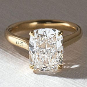 May include: A gold engagement ring featuring a large, cushion-cut diamond. The diamond is held in place by four prongs and has a sparkling, multifaceted surface. The ring's band is plain, polished gold. The text "ELOYA JEWEL" is visible.