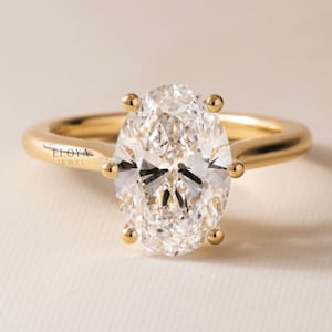 May include: A gold engagement ring with a large, oval-cut diamond. The diamond is held in place by four gold prongs. The ring band is a smooth, polished gold. The brand name "ELOYA JEWEL" is printed on the ring.