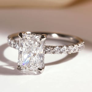 May include: A radiant-cut diamond ring with a pavé band. The large, rectangular diamond is the focal point, set in a four-prong setting. The band is adorned with smaller diamonds, adding sparkle. The ring is set on a white surface.